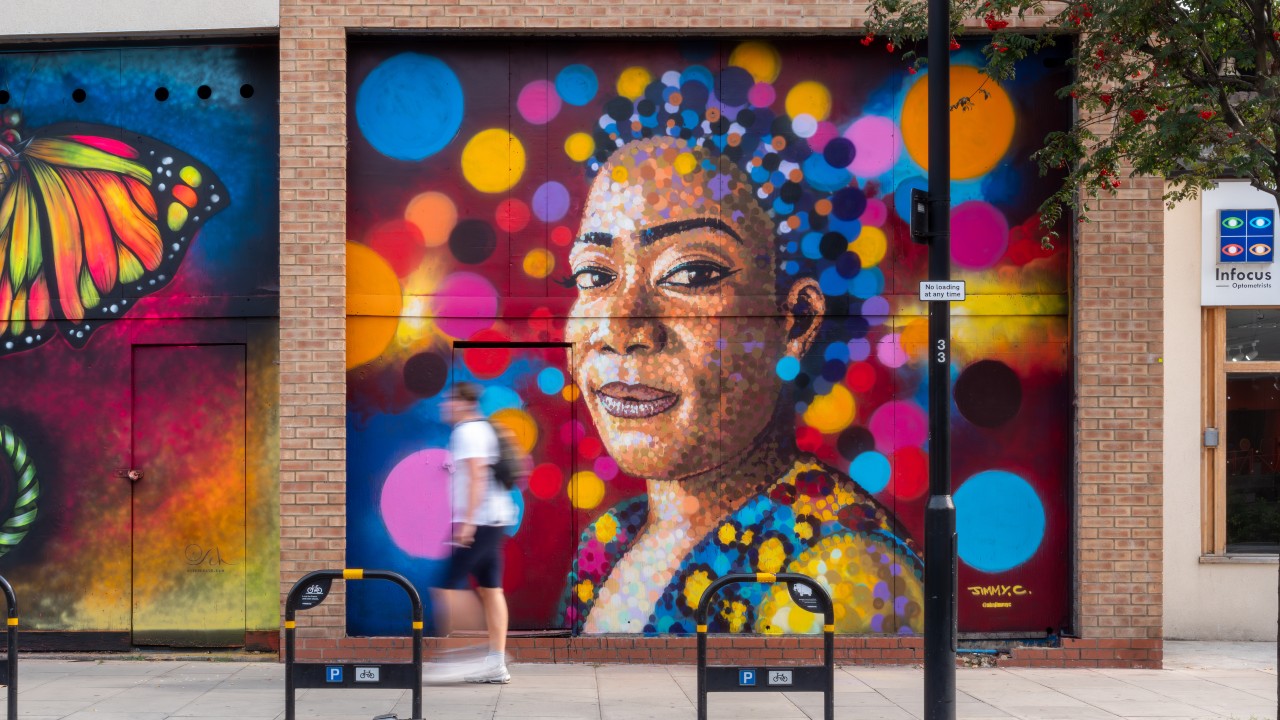 Colourful street art mural featuring a portrait of a black woman with bright dots of colour around her face, including blue, orange and pink splashes of colour, on brick London wall.