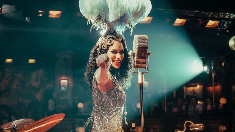 Get ready for an evening of wild Harlem jazz, captivating jazz age tales and spectacular performances at 58th Street. Image courtesy of The Lost Estate. A woman dressed in a silver sparkly outfit stands next to a microphone smiling and pointing to the audience.