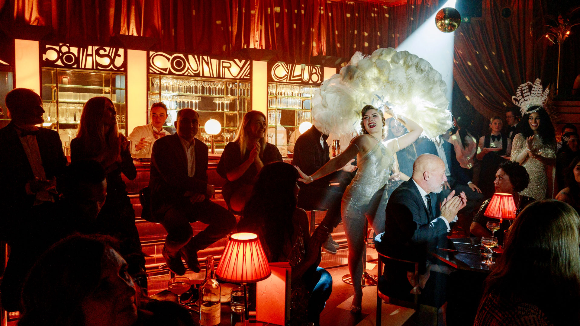 Spend an evening in a 1930s speakeasy filled with glitz and glamour at 58th Street. Credit: The Lost Estate. Image courtesy of The Lost Estate. Audience at The Lost Estate watching a cabaret performance in themed dress under a spotlight with a themed bar in the background