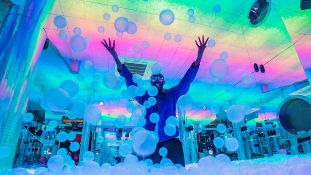 a man stood in the middle of an indoor ball pit throwing balls in the air, with bright colourful lights lighting up the room