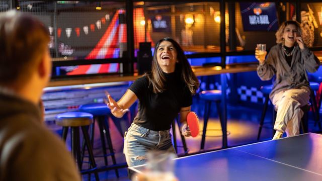 one woman sat on a chair, one in action playing ping pong smiling and the back figure of a man holding a drink watching the ping pong game