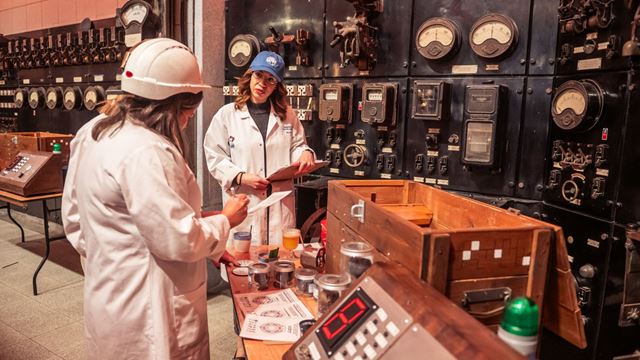 Visitors in lab coats examine samples at a table inside an industrial control room filled with vintage gauges and electrical panels.