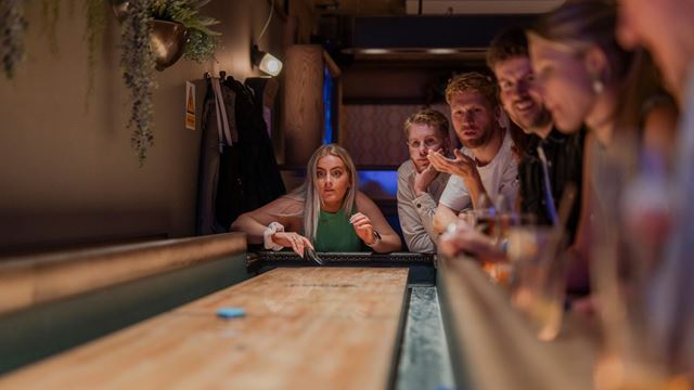 Group of people standing around a shuffleboard table in a dimly lit bar, watching a puck slide across the wooden surface toward the scoring area.