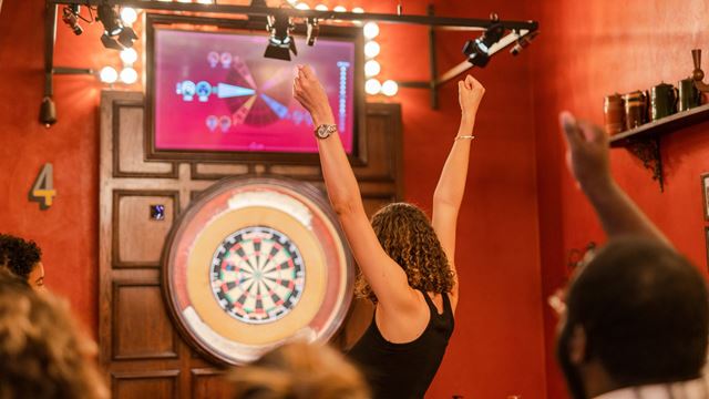 Person standing in front of a dartboard with both arms raised while others watch in a brightly lit indoor bar setting.