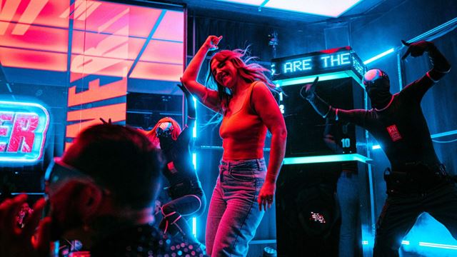 People dancing on a brightly lit stage in a neon-coloured indoor venue, with LED screens, digital displays, and bold blue and pink lighting surrounding the scene.