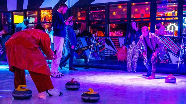 People playing indoor curling on an illuminated rink, with curling stones on the ice and spectators standing nearby in a neon-lit bar and games venue with colourful wall art and large windows in the background.