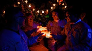 four people wearing jumpsuits, sat around a table and smiling, holding light-up drinks in a dark leafy space with fairy lights in the background