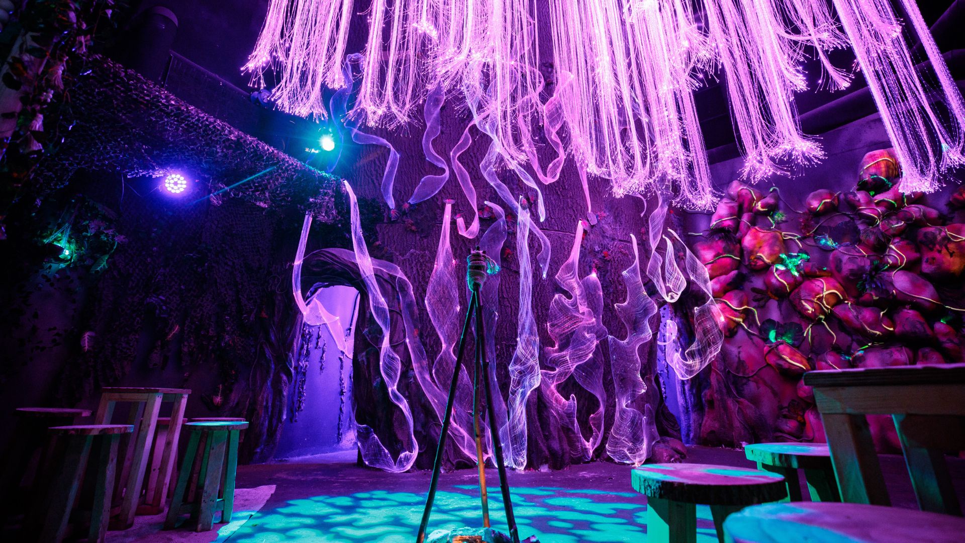 Explore immersive theatrical sets and interact with the actors in Avora: New World Cocktail Experience. Image courtesy of Golden Tours. low angle shot of an immersive mystical dark room, with vibrant pink, purple and blue lights and hanging lights