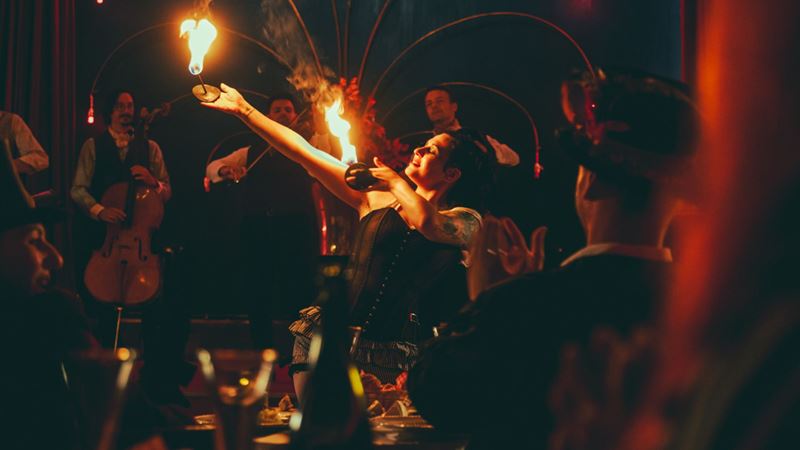 Feel the heat of the night with daring fire acts and spellbinding artistry at CHAT NOIR. Image courtesy of The Lost Estate. A performer captivating the audience with a fire act while musicians play in the background at CHAT NOIR