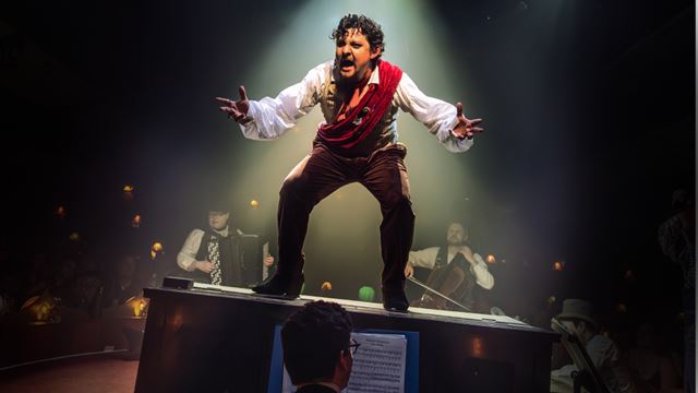 The actor playing the impresario Rodolphe Salis is standing on top of a piano and raising his arms towards the audience at the CHAT NOIR!