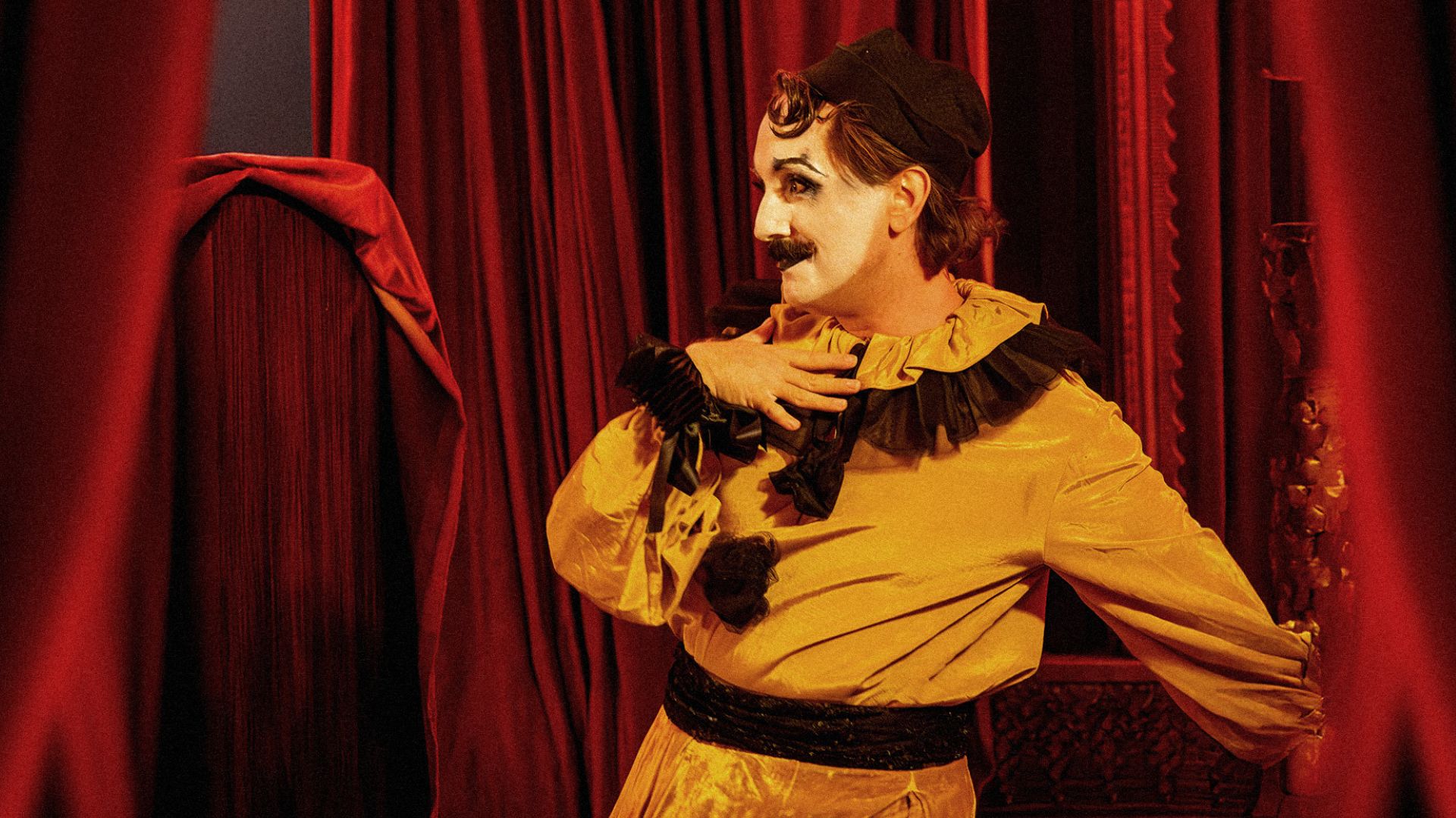 A performer in a striking yellow costume with black accents posing dramatically against deep red velvet curtains at CHAT NOIR