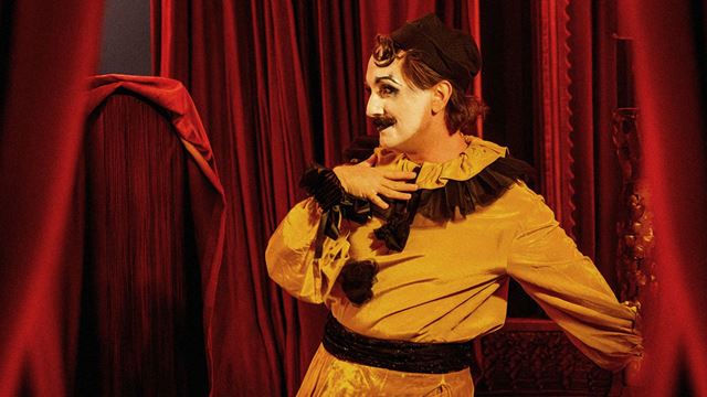 A performer in a striking yellow costume with black accents posing dramatically against deep red velvet curtains at CHAT NOIR