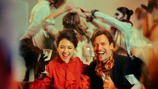 Two guests dressed in vintage-inspired attire enjoying cocktails, with lively dancers performing in the background at CHAT NOIR.