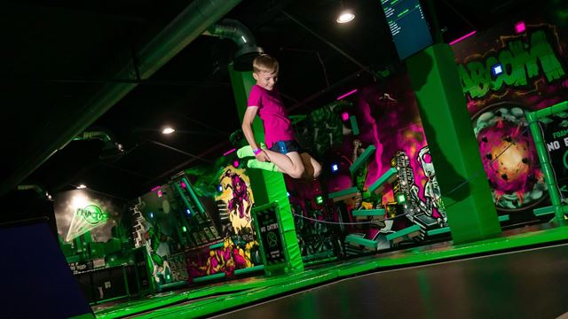 A child balances on an obstacle above a trampoline area at Flip Out, surrounded by colourful neon lights and adventure-course challenges.