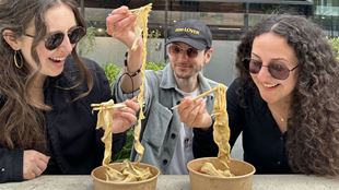Group of three sat at a table smiling and eating a bowl of noodles during an outdoor food treasure hunt experience in London.