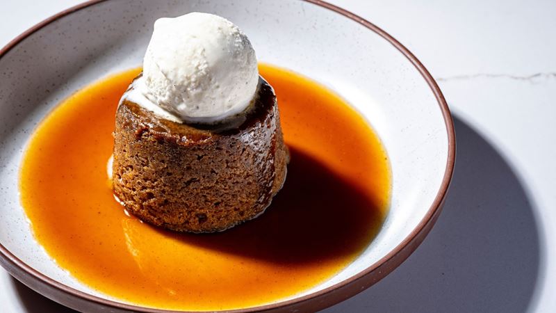 Hell's Kitchen London promises exsquisite cuisuine alongside a memorable immersive dining experience. Image courtesy of Gordon Ramsay Brand. A white plate with a sponge pudding and a ball of white ice cream at Hell's Kitchen London.