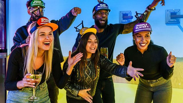 Five people wearing brightly coloured ‘player’ caps cheer and gesture excitedly while playing an immersive group game in a neon-lit room.