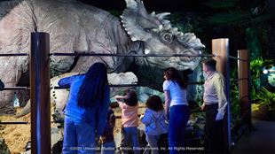 Visitors, including children and adults, observe a life-sized animatronic Triceratops in a jungle-themed exhibit, enclosed by safety railings. The dinosaur features realistic textures and is part of the Jurassic World: The Experience