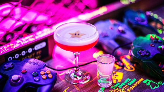 Bright pink cocktail on a table surrounded by retro gaming controllers in a neon-lit bar.