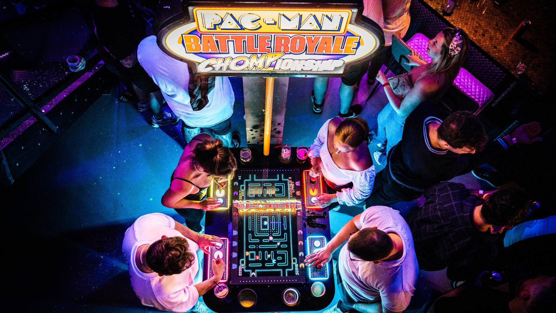 People gathered around a Pac-Man arcade table in a colourful, neon-lit gaming bar.