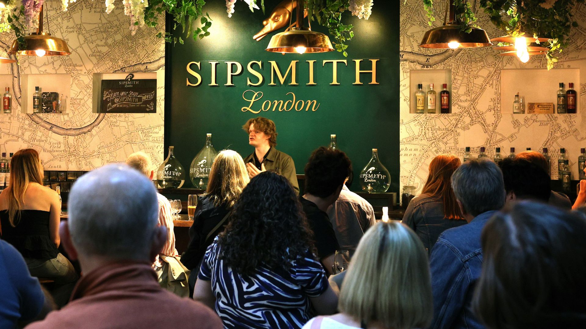 Guests seated at a Sipsmith tasting session while a host presents in front of a Sipsmith London display.