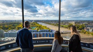 Three people standing at a panoramic viewing platform overlooking the River Thames, Battersea Power Station, and surrounding cityscape under a partly cloudy sky.