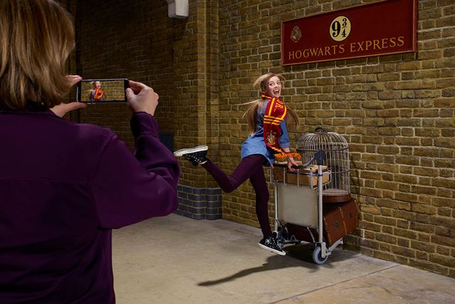 A young woman with long hair is posing pushing a Harry Potter trolley, half way trough a wall.