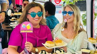 two women sat at a table with afternoon tea, smiling and wearing joke-glasses and holding funny signs and smiling