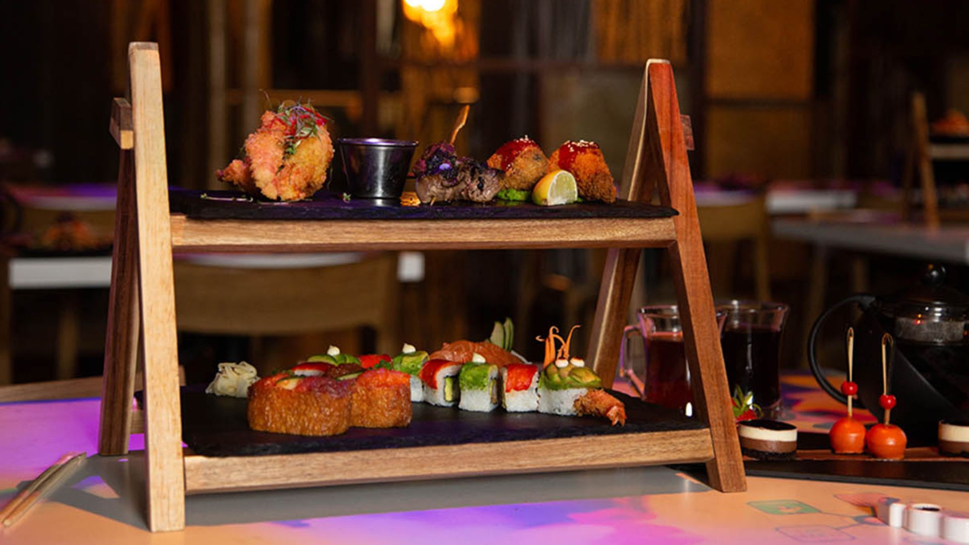Two-tier wooden serving stand displaying sushi rolls, fried tempura and small Japanese-style dishes on a restaurant table with low lighting.