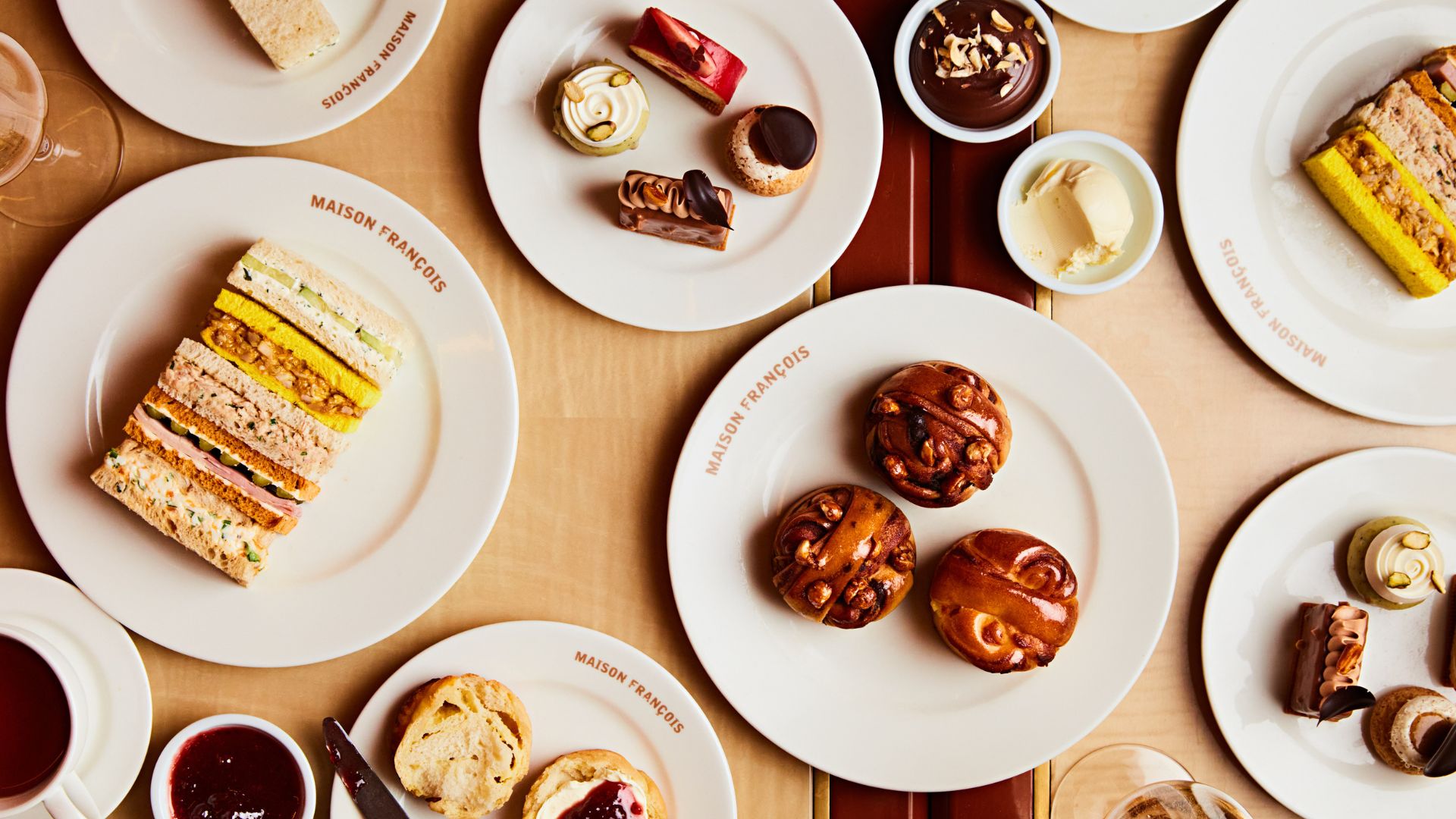 Overhead view of a Maison François afternoon tea featuring finger sandwiches, babkas with jam and cream, pastries and chocolate desserts served on branded white plates.