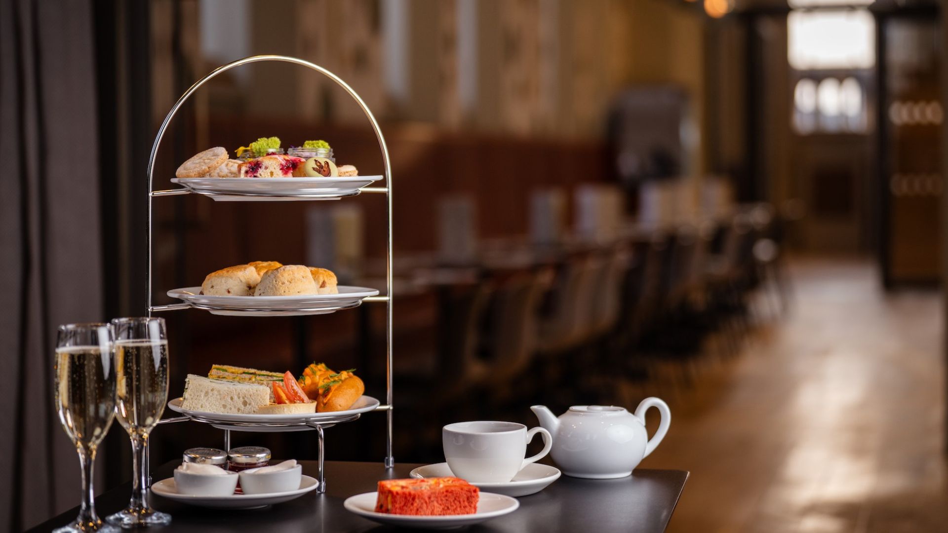 Three-tier afternoon tea stand with finger sandwiches, scones and cakes on a table alongside a teapot, teacup and two glasses of Champagne in a restaurant setting.