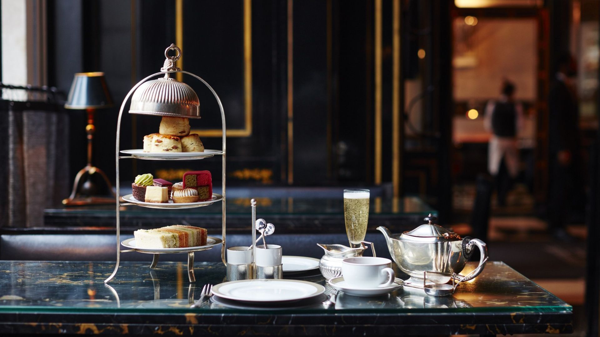 Three-tier afternoon tea stand with scones, cakes, and finger sandwiches on a marble table, alongside a silver teapot, teacup, and a glass of sparkling wine in an elegant, softly lit dining room.