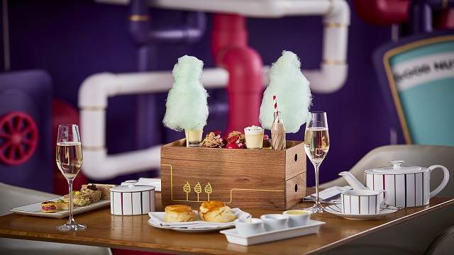 A dining table with display boxes full of cakes and candy floss, scones on plates, glasses of champagne and a teapot, cup and saucers
