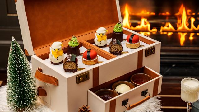 Festive afternoon tea display in a cream and tan leather chest, featuring Christmas-themed pastries shaped like Santa and trees, chocolate yule logs, red and black mousse cakes, and scones with jam and cream, set on a fluffy white surface with a miniature Christmas tree and a roaring fireplace in the background.