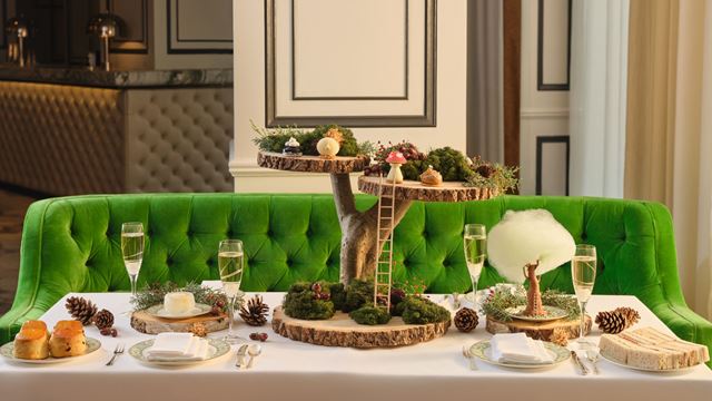 A whimsical woodland-themed afternoon tea display set on a white table with a bright green velvet banquette. The centerpiece is a two-tier stand made of rustic wooden slabs and moss, decorated with miniature mushrooms and a tiny ladder. Plates of scones, pastries, and desserts surround the stand, alongside champagne flutes and pinecones for a festive touch.