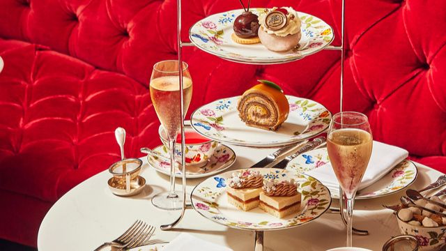 Ein Tisch mit weißem Tischtuch und Etagere auf der ein festlicher Nachmittagstee serviert wird, vor einem leuchtend roten Samtsofa.