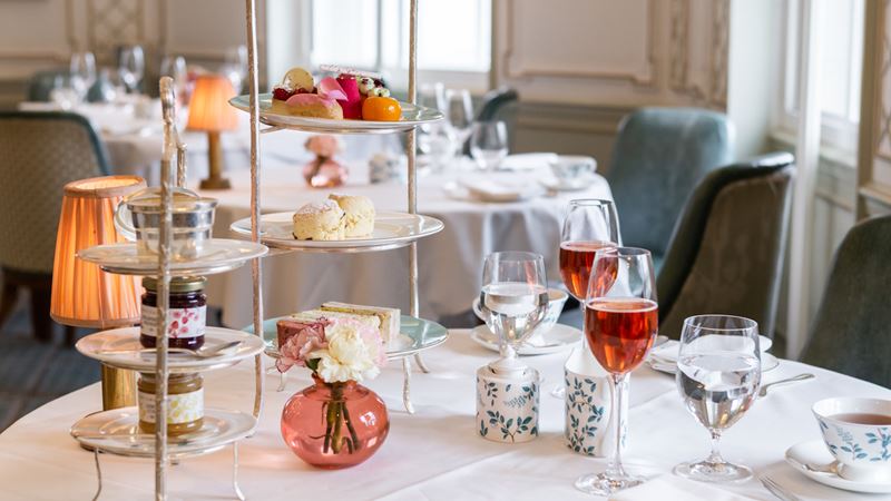 Champagne afternoon tea at The Diamond Jubilee Tea Salon. Credit: Fortnum & Mason. Image courtesy of Fortnum & Mason. Afternoon tea cakes, scones and sandwiches in a three-tiered tray, with preserves, glasses of champagne and cups of tea on a table with white tablecloth.
