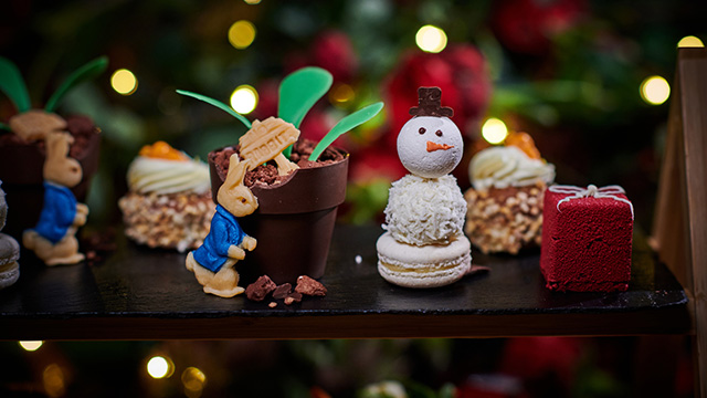 Kleine Kuchen, unter anderem in Form von Peter Rabbit, einem Schneemann und einem Würfel, auf einer Holzplatte, mit Weihnachtslichtern im Hintergrund.