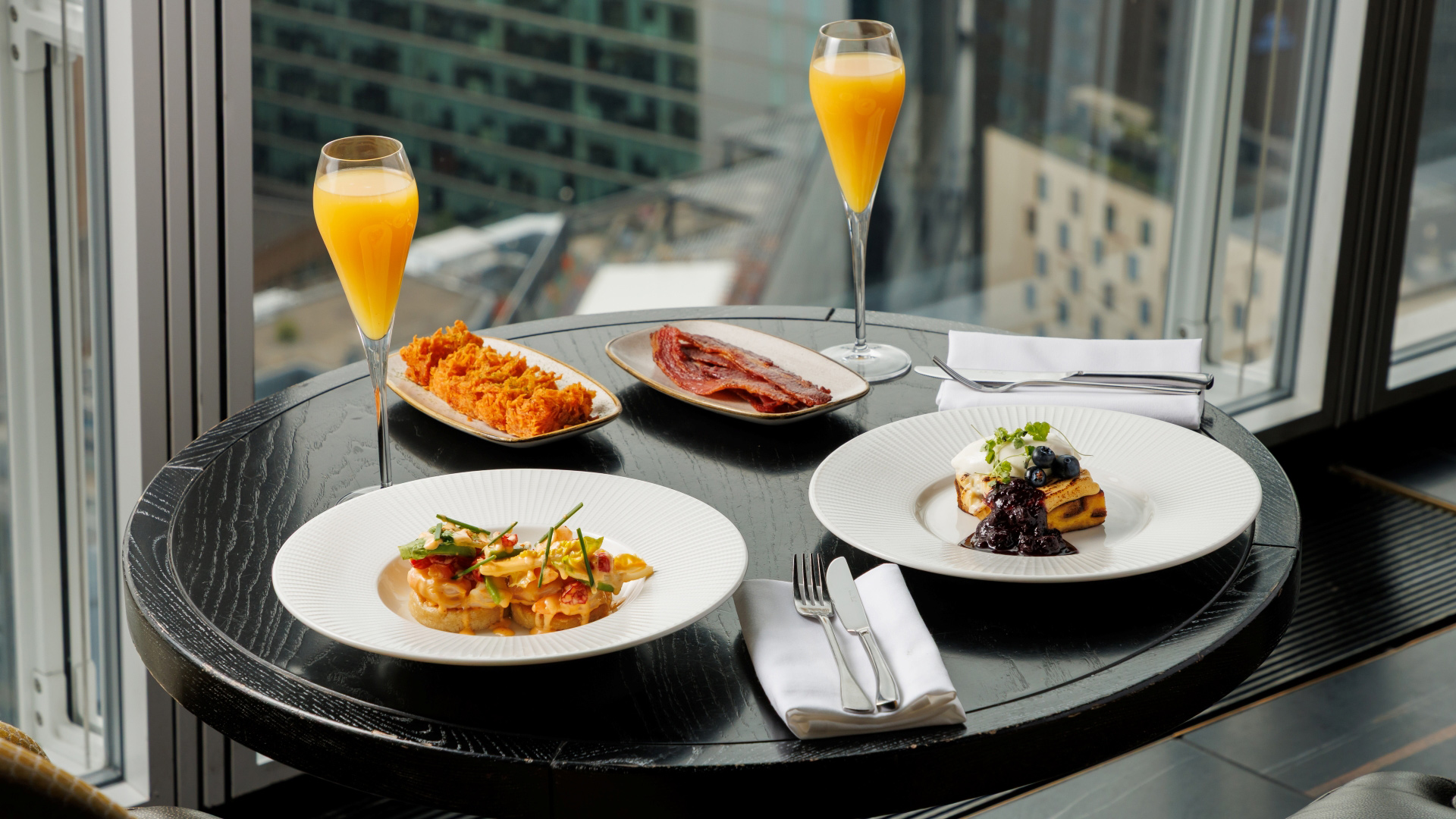 A photo of a table at Aqua Shard filled with brunch dishes and two flutes with mimosas with a background of a view over London through floor-to-ceiling windows