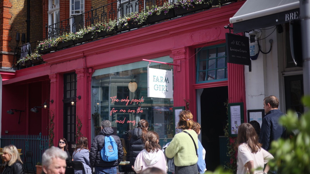 A photo of the exterior of Farm Girl painted in a berry pink colour with people stood outside and neon lights in the window that say 