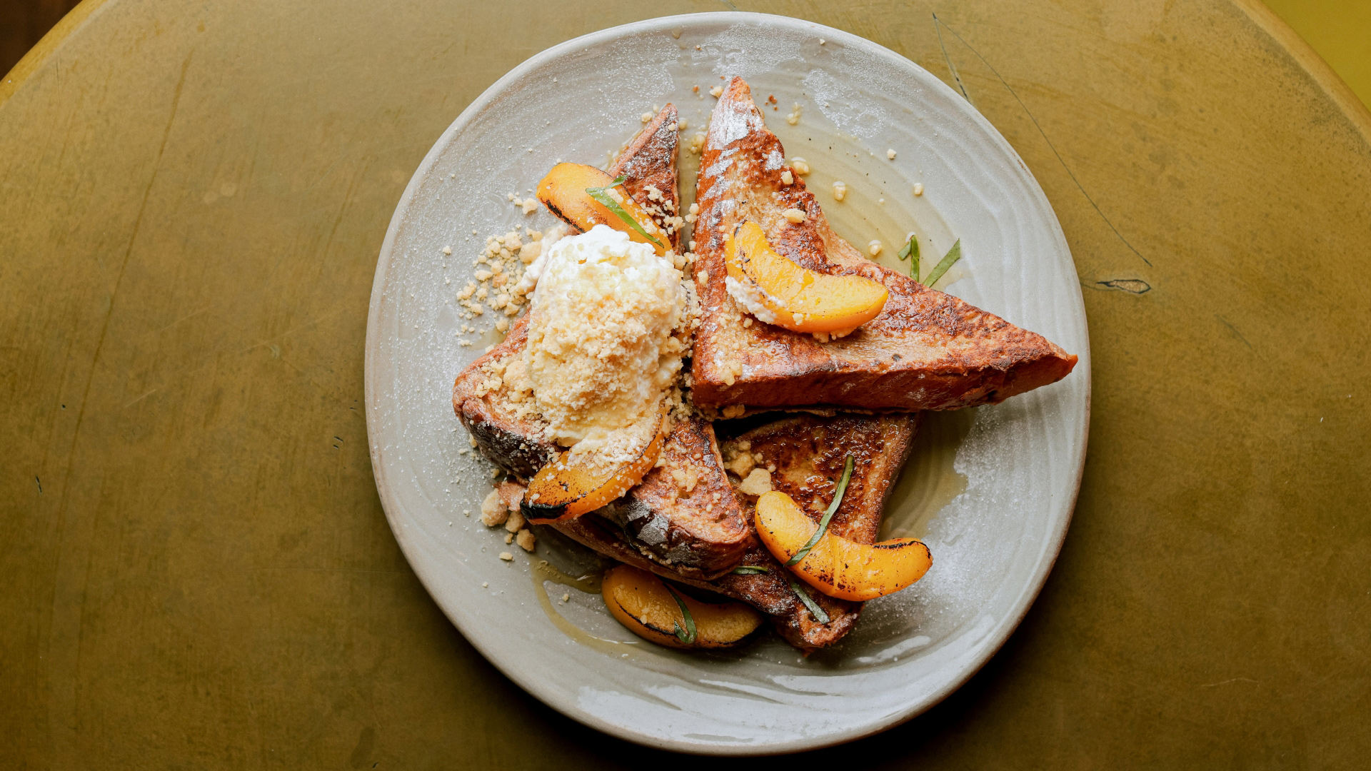 A bird's-eye photo of a plate of french toast topped with a dollop of mascarpone, burnt peach slices, basil and a peach maple syrup