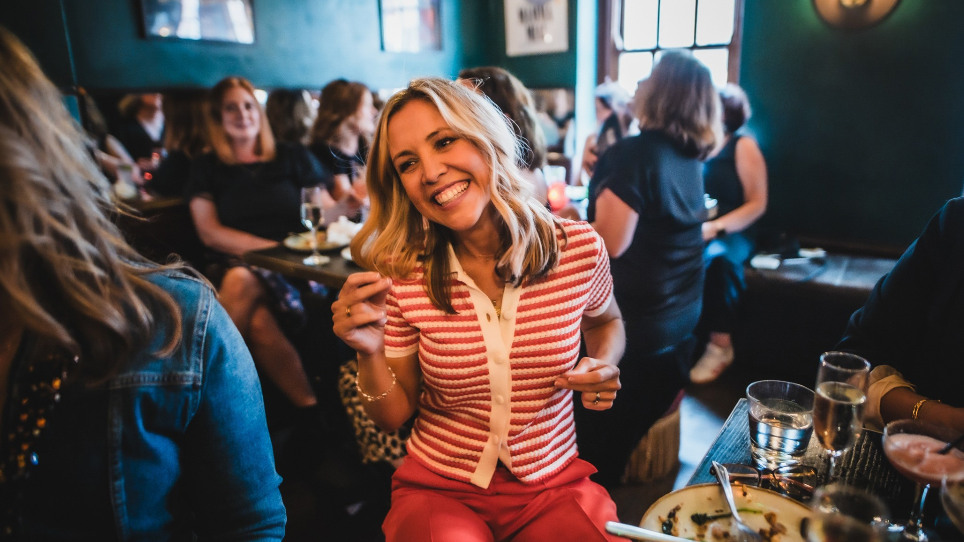A photo of a woman in a striped top smiling and clicking her fingers while dancing at her table with people in the background and an empty plate of food in front of her