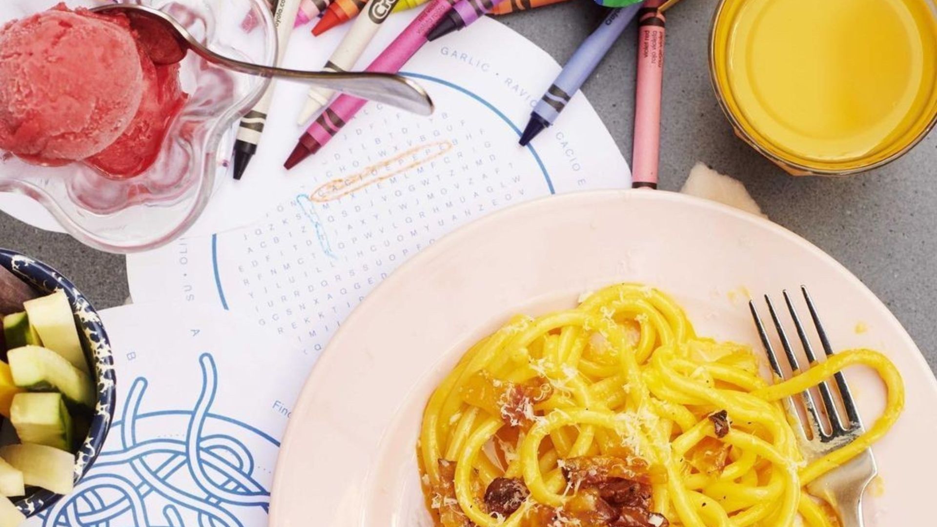 A bowl of spaghetti pasta with grated cheese on a table beside crayons, a colouring sheet, fruit pieces and a dessert dish.