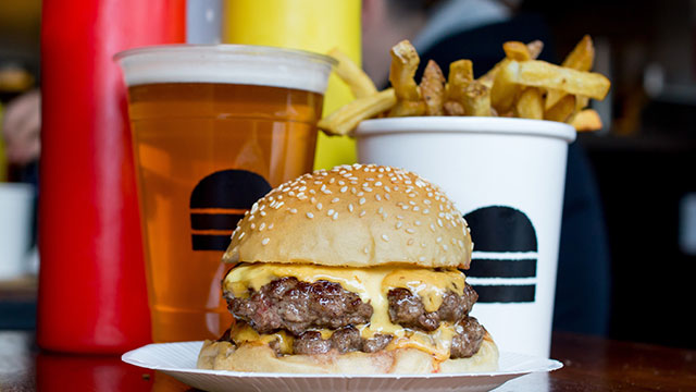 A burger on a paper plate with a beer, chips and bottles of ketchup and mustard behind