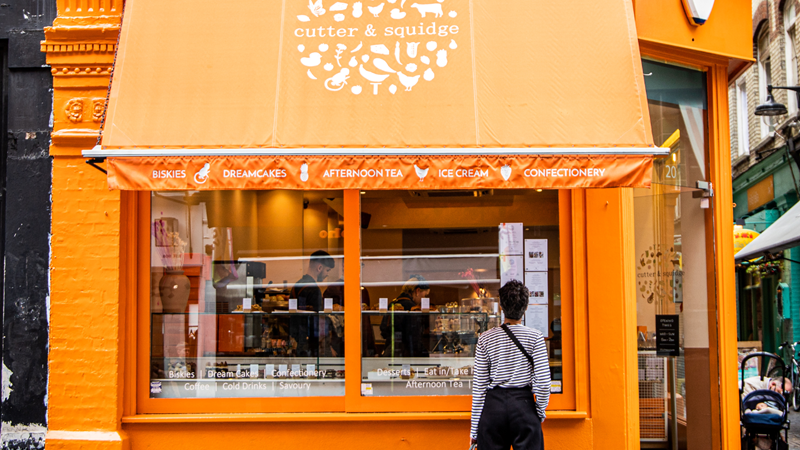 Head to the Cutter and Squidge shop in Soho to satisfy your sweet treat cravings. Credit: Cutter & Squidge. Image courtesy of Cutter & Squidge. A woman in jeans and a striped top stands outside the bright orange Cutter and Squidge shop on the corner of a street in Soho with baked goods in the window