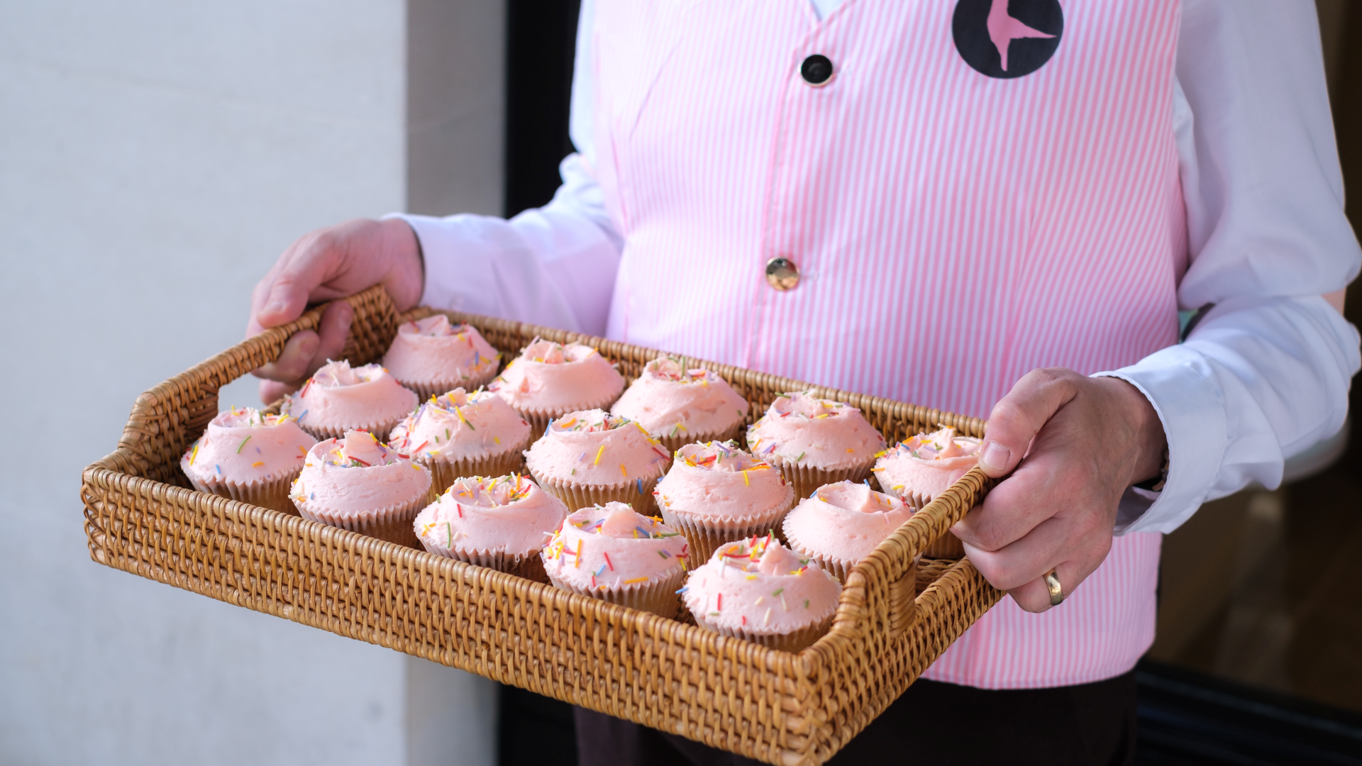 A Hummingbird Bakery employee wearing a white shirt and pink waistcoat holds a wooden tray with pink cupcakes topped with sprinkles