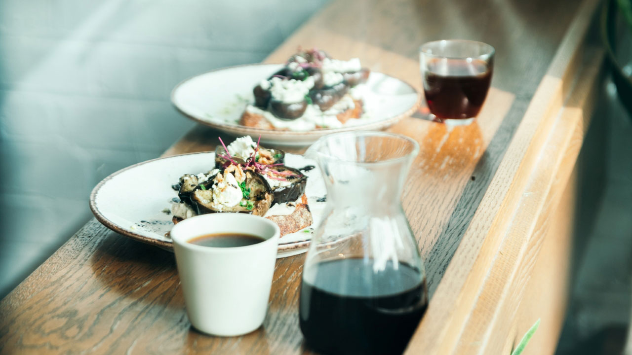 A photo of two plates of brunch dishes, a mug of coffee and a glass of coffee with a carafe filled with black coffee on a wooden table inside Prufrock Coffee