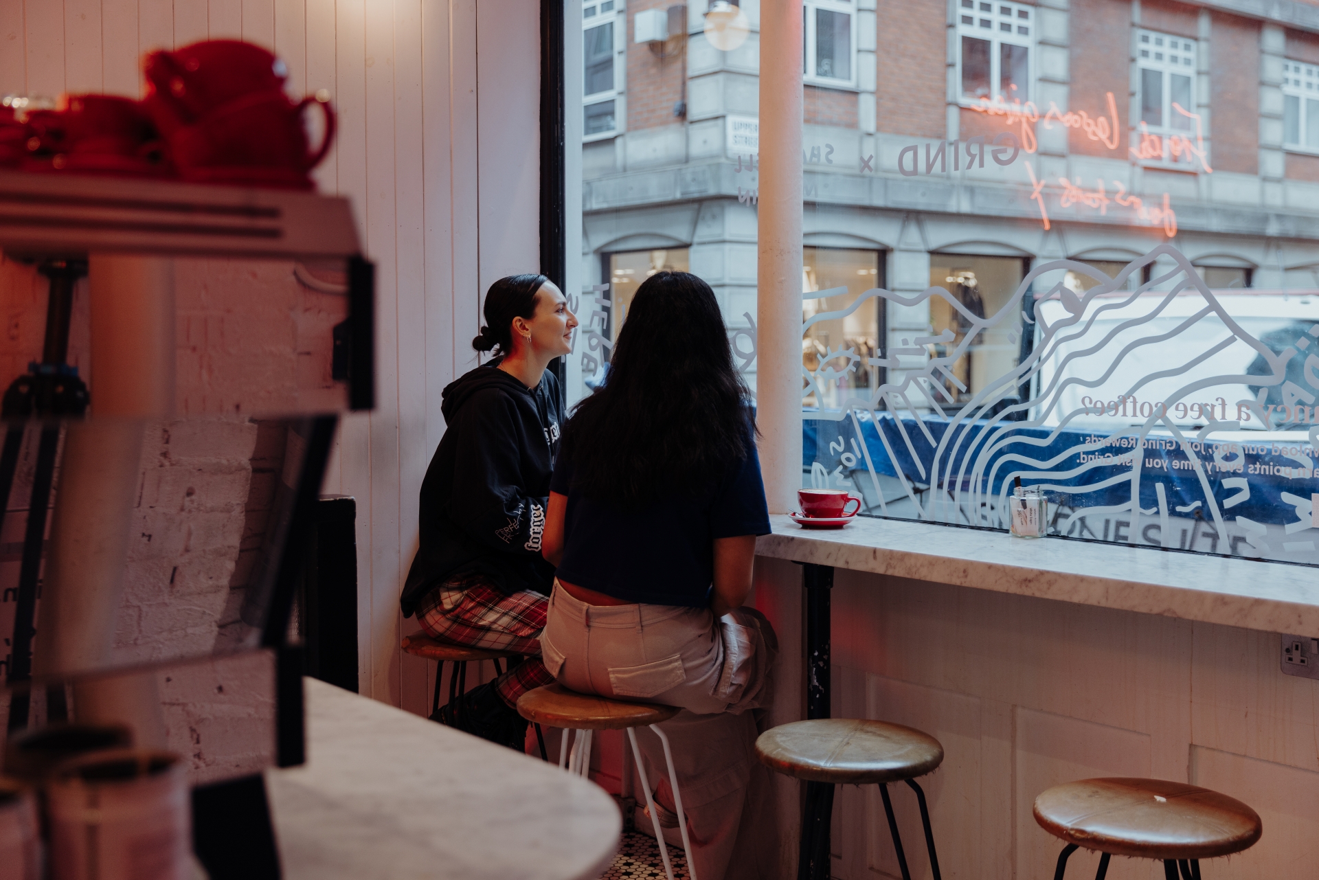 A photo of two friends sat on stools inside Soho Grind coffee shop looking out the window onto the street with a coffee machine and neon lights in the background