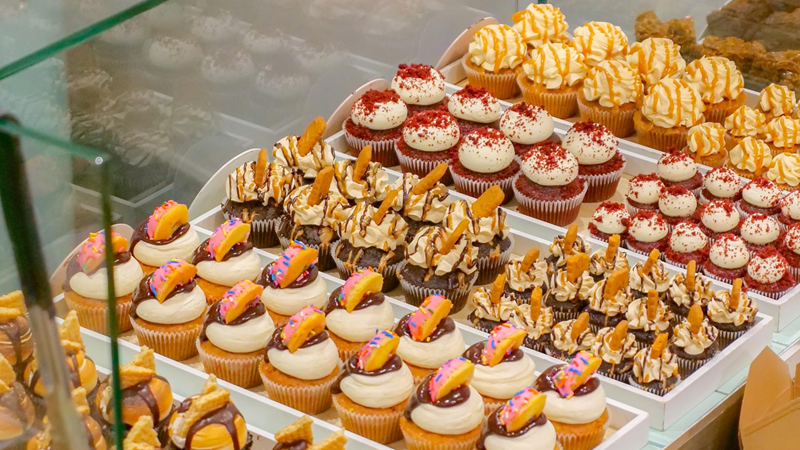 Choose from a mouthwatering selection of fresh cakes at Crumbs and Doilies. Credit: Crumbs and Doilies Ltd. Image courtesy of Crumbs and Doilies. A selection of cupcakes on the counter neatly lined up behind a glass wall each with a generous dollop of icing and decorations