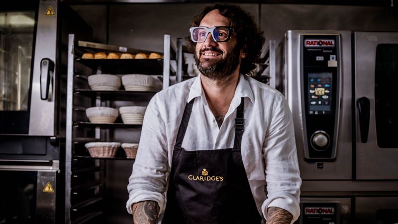 Richard Hart at work at Claridge’s Bakery, bringing classic British bakes to the iconic Mayfair hotel from January 2026. Image courtesy of MAYBOURNE. Baker Richard Hart wearing a Claridge’s apron inside a professional bakery kitchen with bread racks and ovens behind him.